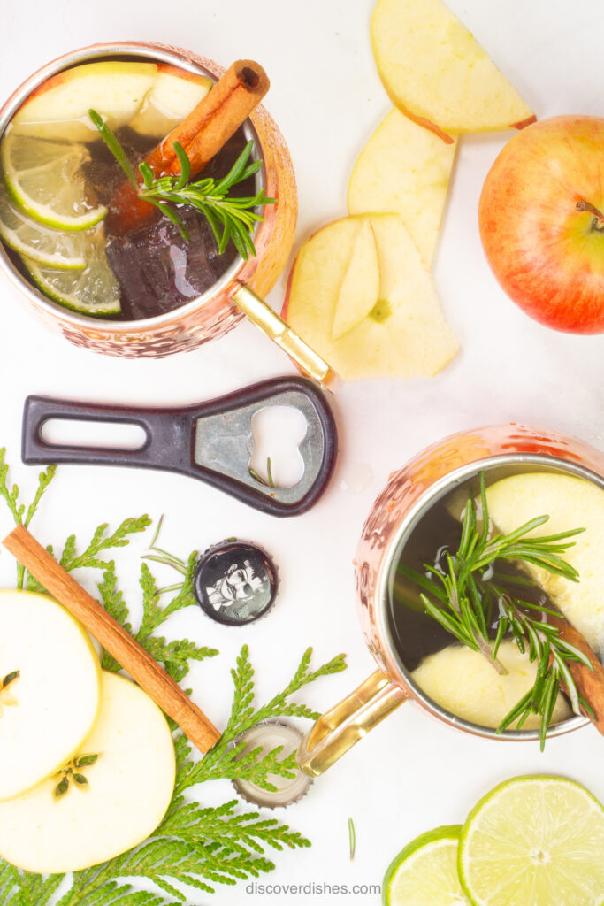 A bottle opener and apple slices arranged around two apple cider moscow mules.