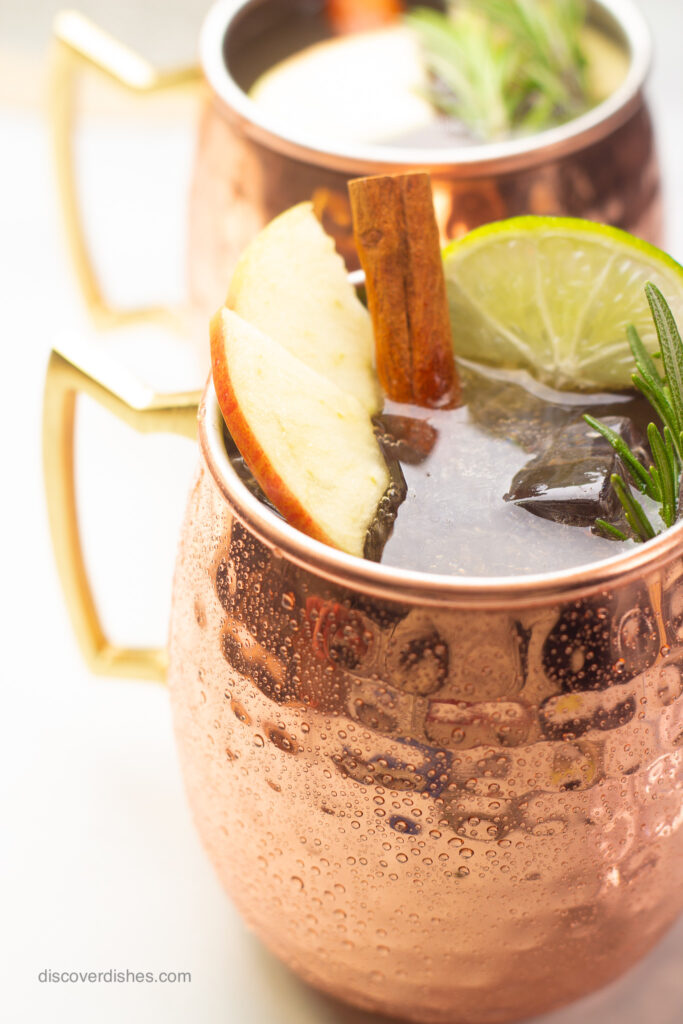 An apple cider moscow mule garnished with apples, limes, cinnamon sticks, and rosemary.