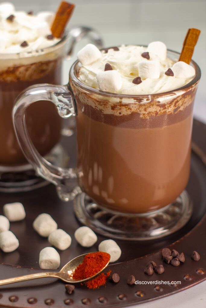 A cup of mexican hot chocolate with a spoon of cayenne pepper next to it.